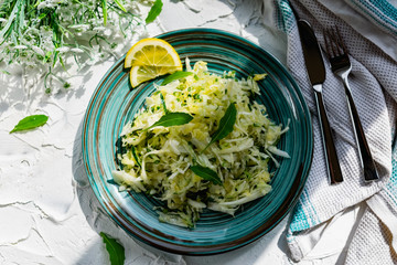 Crispy salad of fresh vegetables with herbs. Young cabbage with cucumber and arugula leaves, decorated with lemon and a bouquet of white flowers on a light textured stone table with Cutlery