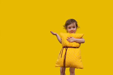 a beautiful little girl with a yellow pillow, looking at the camera, smiling and holding a comfortable pillow on a yellow background with one hand, the other hand holding her hand up.