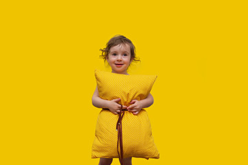 a beautiful little girl with a yellow pillow, looking at the camera laughing and hugging a pillow on a yellow background.  Concept of comfortable pillows for everyone