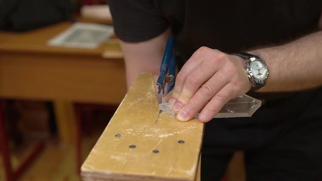 master holds transparent plexiglass on brown wooden support and cuts with blue metal fretsaw in workshop close view