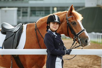 Beautiful professional female jockey standing near horse. woman horse rider is preparing to...
