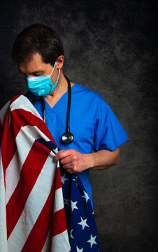 Sad/pensive male doctor in blue hospital scrubs with face mask and stethoscope, nursing the Stars & Stripes American flag close to his chest, and standing against a dark studio background.