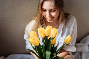 Beautiful young blond woman with tulip bouquet. Close-up spring portrait. Happy and romantic woman at home interior with sun rays and a bouquet of yellow flowers. Girl in underwear and white shirt