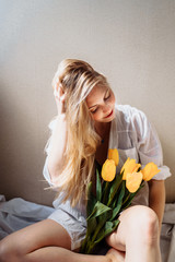 Vertical photo. Attractive blonde girl in lingerie and white shirt sitting in sunshine. Holding yellow tulips, sniffing them, hiding behind flowers, enjoying the sun and life, smiling, laughing