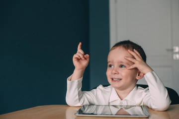 Cute little boy with big blue eyes in a white T-shirt sits at a table with a tablet and shows thumb up. Self isolated. Online education. Home schooling