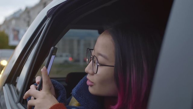 Young cheerful asian woman with pink hair riding in taxi, looking from window and taking picture of city with smartphone