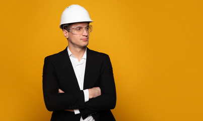 Serious business. Close-up photo of a young architect in formal outfit and hard hat, who is standing with folded arms and looking aside.