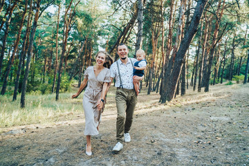 Happy young family having fun running through the evening summer forest. Dad holding in his hands a one-year-old child boy. Parents happily bouncing and having a good time out of town