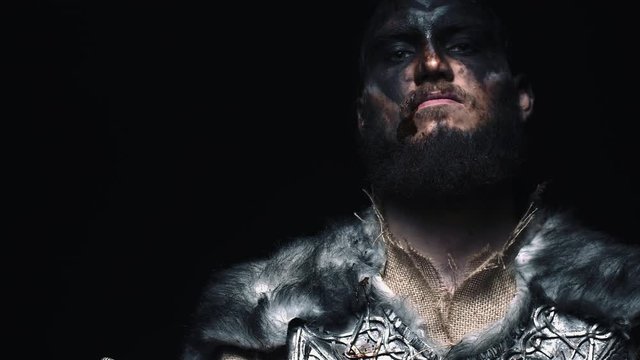 The Viking man looks into the camera and slowly lowers his head, the warrior holding the axe next to his face. Viking in war paint