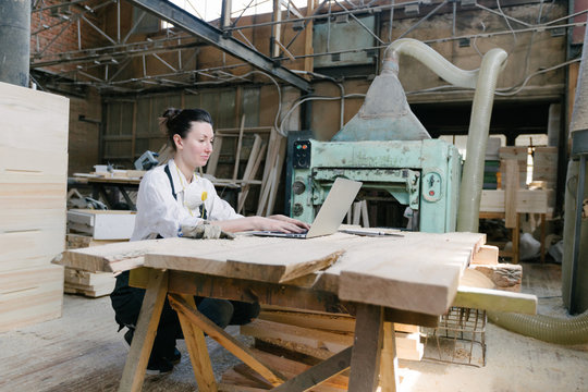 Confident Woman Working As Carpenter In Her Own Woodshop. She Using A Laptop And Writes Notes While Being In Her Workspace. Small Business Concept.