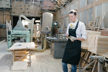 Confident woman working as carpenter in her own woodshop. She using a tablet pc and writes notes while being in her workspace. Small business concept.
