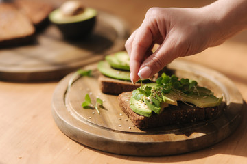 Woman put mustard seedlings on avocado sandwich with dark rye toasted bread made with fresh sliced avocados from above with sesame seeds