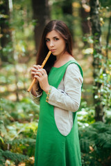 Woman in a green medieval dress plays the wooden flute while standing in a grove against the background of pines. Girl in a gloomy forest plays music on a handmade pipe. Young witch in a gloomy forest © Михаил Гута