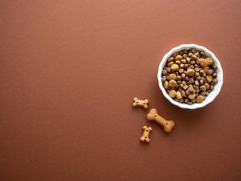 Hunde Trockenfutter in einem wei&szlig;en Futternapf  und Leckerlie auf einem braunen Hintergrund, Draufsicht, Tierthema