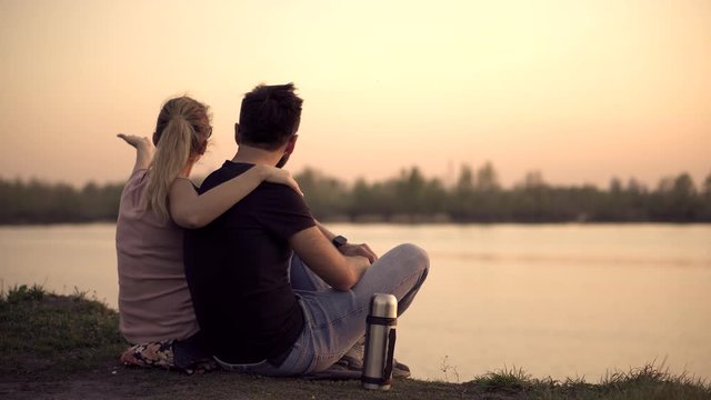 Romantic Couple Sitting By The River With Cups Of Tea. Couple Relaxing End Watching On Sunset. Couple Is Looking At Red Sunset.	