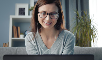 Close-up of young beauty wearing earphones and spectacles sitting on couch looking at laptop screen - obrazy, fototapety, plakaty
