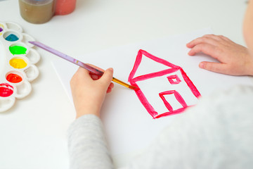 Child is drawing red house with watercolors on the white sheet of paper.