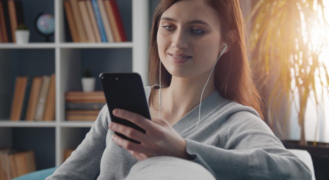 Casual Young Female Using Mobile Phone And Earphones Leaning Sideways On Sofa In Apartment