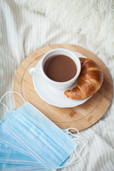 A cup of hot cocoa, a croissant, a wooden stand, a medical mask on the bed. Quarantine time. Homeliness.