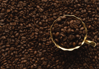 Golden cup on saucer filled with coffee beans on blurred background full of coffee beans