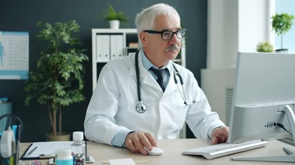 Serious serior man doctor is using computer typing sitting at desk in hospital office working alone. Modern technology, profession and medicine concept. - Powered by Adobe