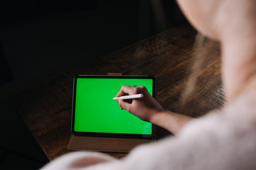 Young woman using black tablet device with green screen. Woman holding tablet, scrolling pages while sitting on the couch in the living room. Chroma key. View over shoulder. Close up