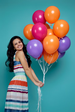 Smiling carefree curly girl in dress holding bright colorful air balloons isolated on blue background. Beautiful happy young woman on a birthday party.