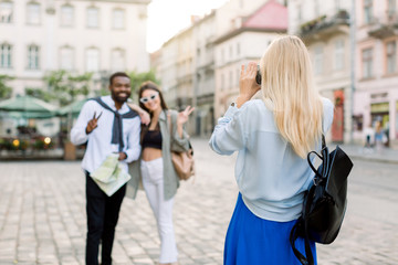 Fototapeta premium Summer holidays, technology and city travel concept. Back view of pretty Caucasian blond girl taking photo of two multiethnic friends, dark skinned man and Caucasian woman in the city