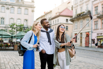 Multiethnic friends, tourists, young African man and two Caucasian women, wearing casual wear is exploring new city together, smiling and making photo on smartphone