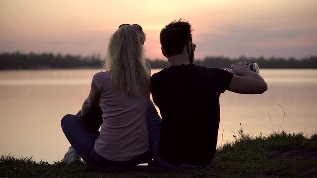 Romantic Couple Sitting By The River With Cups Of Tea. Couple Relaxing End Watching On Sunset. Couple Is Looking At Red Sunset.