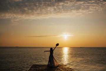 Silhouette of young woman in long dress watching sea sunset. Breathtaking nature background.  Photo with copy space. Woman with hands up enjoying beautiful sunset. 