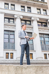 Young European Businessman traveling, working in New York City, wearing gray blazer, white shirt, tie, black pants, standing on street outside vintage office building, working on laptop computer..