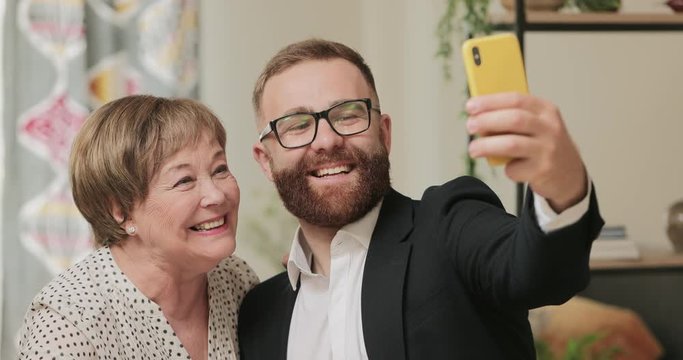 Handsome man in suit doing selfie with his cheerful grandmother while visiting her at home. Mature woman in 70s kissing his adult grandson and possing to photo. Concept of fun and family.