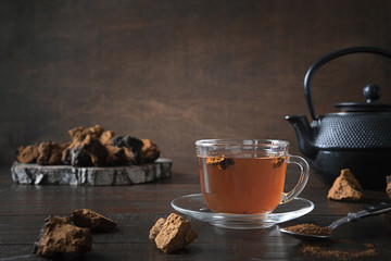 Chaga mushrooms in glass cup and teapot. Trendy healthy infusion from birch on dark wooden background. Close up.