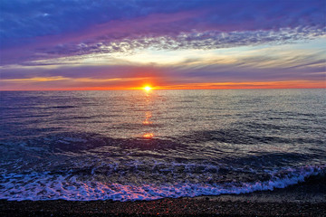 Sunset over the Black Sea. The sun was almost hidden and painted the clouds pink. Lacy foam of waves and bright patches of light on pebbles of the shore. Bright sunny walkway on the surface of the sea