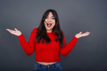 Portrait of beautiful female looks with excitement at camera, keeps hands raised over head, notices something unexpected, isolated over gray wall. Lovely woman reacts on sudden news.