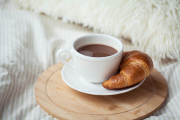 A cup of hot cocoa, a croissant, a wooden stand on the bed. Quarantine time. Homeliness.
