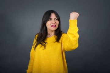 Fierce confident European dark-haired woman holding fist in front of her as if is ready for fight or challenge, screaming and having aggressive expression on face. Isolated over gray background.