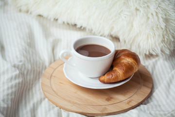 A cup of hot cocoa, a croissant, a wooden stand on the bed. Quarantine time. Homeliness.