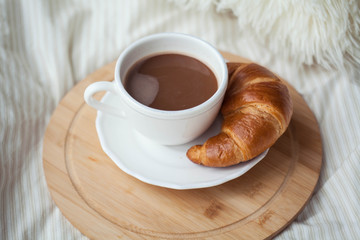 A cup of hot cocoa, a croissant, a wooden stand on the bed. Quarantine time. Homeliness.