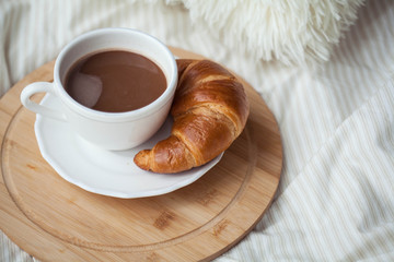 A cup of hot cocoa, a croissant, a wooden stand on the bed. Quarantine time. Homeliness.