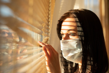 Woman with surgical mask looking out of home window, concept of quarantine during viral pandemic...