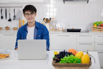 Asian freelance young man working from home with breakfast bread and coffee using laptop in white kitchen