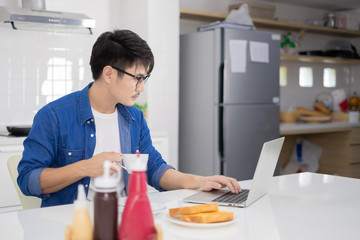 Asian freelance young man working from home with breakfast bread and coffee using laptop in white kitchen