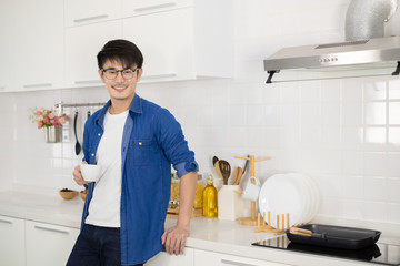 Asian man holding a cup of coffee and sitting in white kitchen.