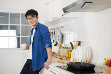 Asian man holding a cup of coffee and sitting in white kitchen.