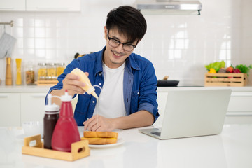 Asian freelance young man working from home with breakfast bread and coffee using laptop in white kitchen