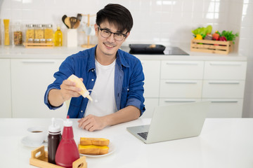 Asian freelance young man working from home with breakfast bread and coffee using laptop in white kitchen