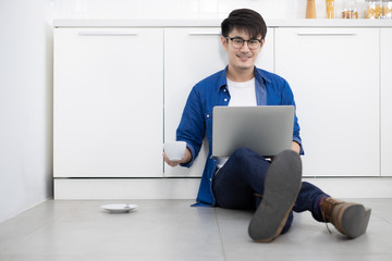 Asian freelance young man working from home with coffee using laptop sittng on the floor in white kitchen