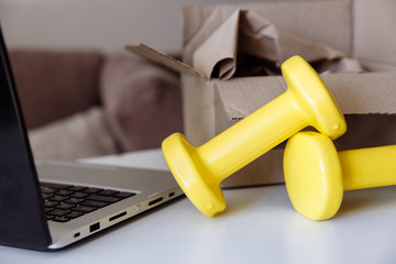 Two yellow dumbbells lie on a white table. Next to it is a silver laptop. In the background is a delivery box.